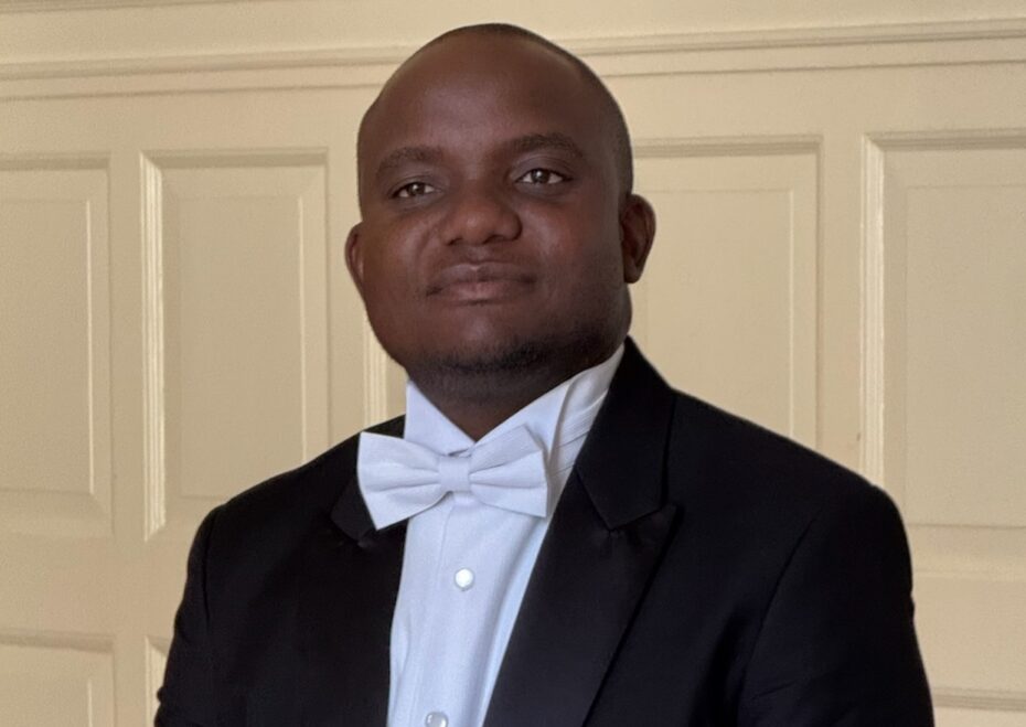 Portrait of a man in a black tuxedo and white bow tie, indoors against cream paneled doors.