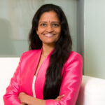Dr Jayasree K. Iyer - OHE Smiling woman with long dark hair wearing a pink blazer sits on a white couch in an office setting.