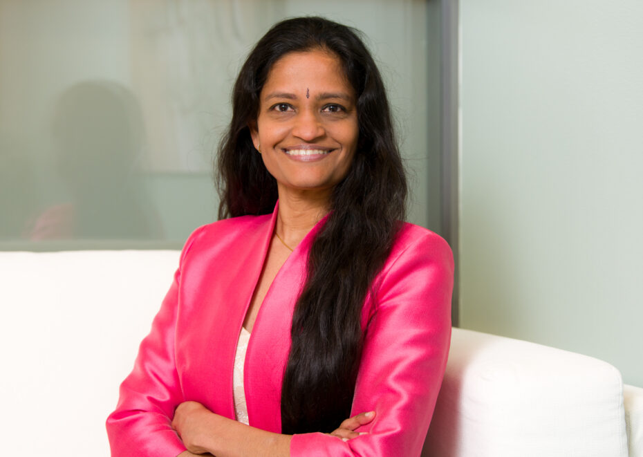 Smiling woman with long dark hair wearing a pink blazer sits on a white couch in an office setting.