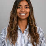 Poushali Ganguli - OHE Smiling woman with long wavy brown hair wearing a blue-and-white striped shirt against a gray background.