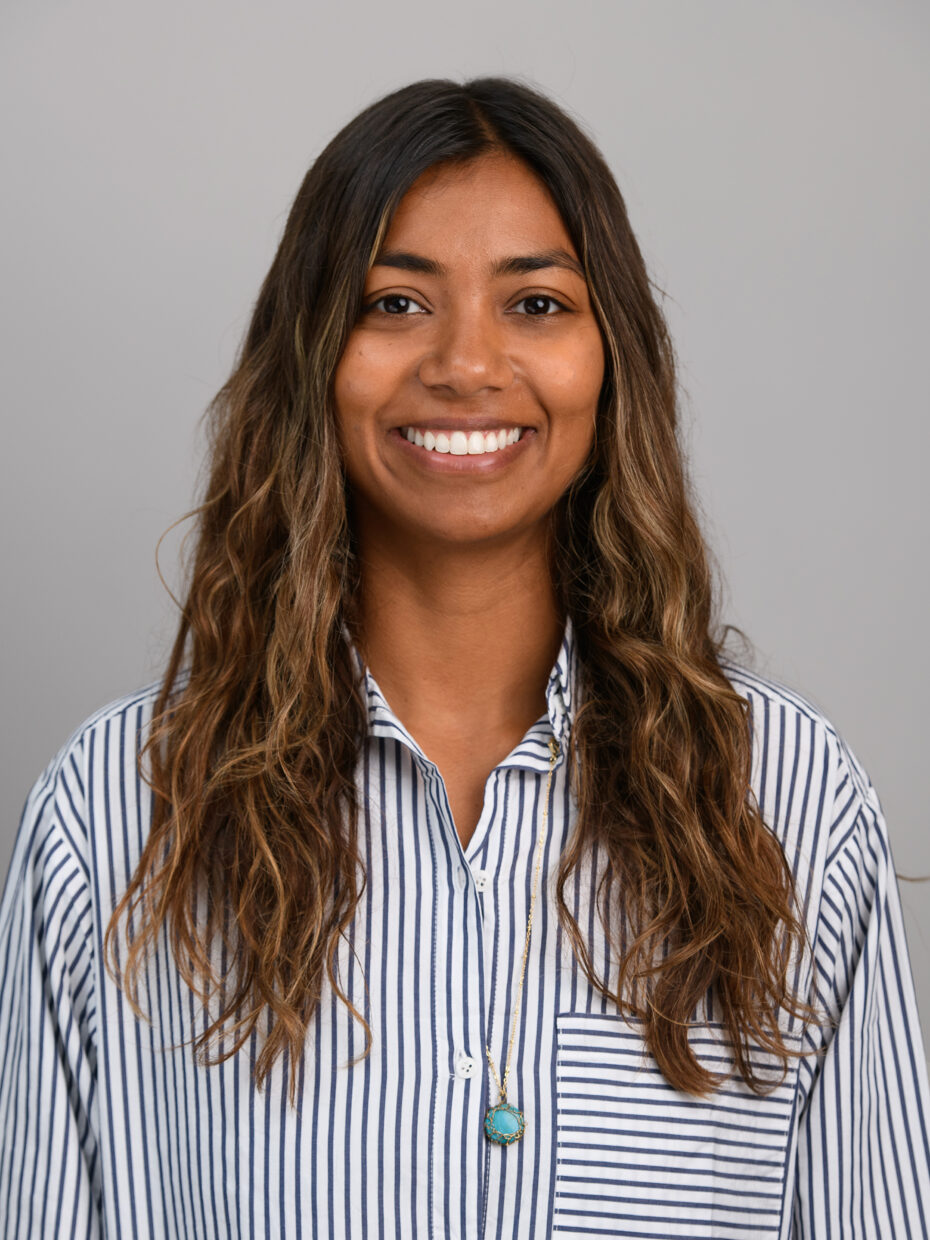 Smiling woman with long wavy brown hair wearing a blue-and-white striped shirt against a gray background.