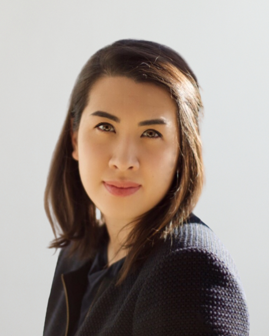 Portrait of a woman with shoulder-length dark hair looking at the camera, wearing a dark textured jacket against a light background.