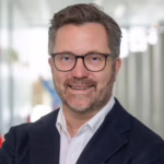 Dr Claus Runge - OHE Smiling man with short gray hair and glasses in a navy blazer and white shirt, professional headshot in an office setting