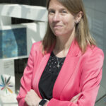 Francesca Colombo - OHE Professional woman with blonde hair in a pink blazer, arms crossed, standing outside a modern building with OCDE signage in the background.