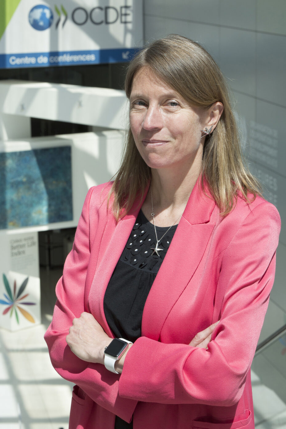 Professional woman with blonde hair in a pink blazer, arms crossed, standing outside a modern building with OCDE signage in the background.