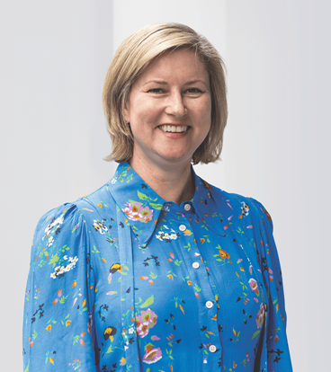 Smiling woman with short blonde hair wearing a blue floral blouse, standing against a light background.