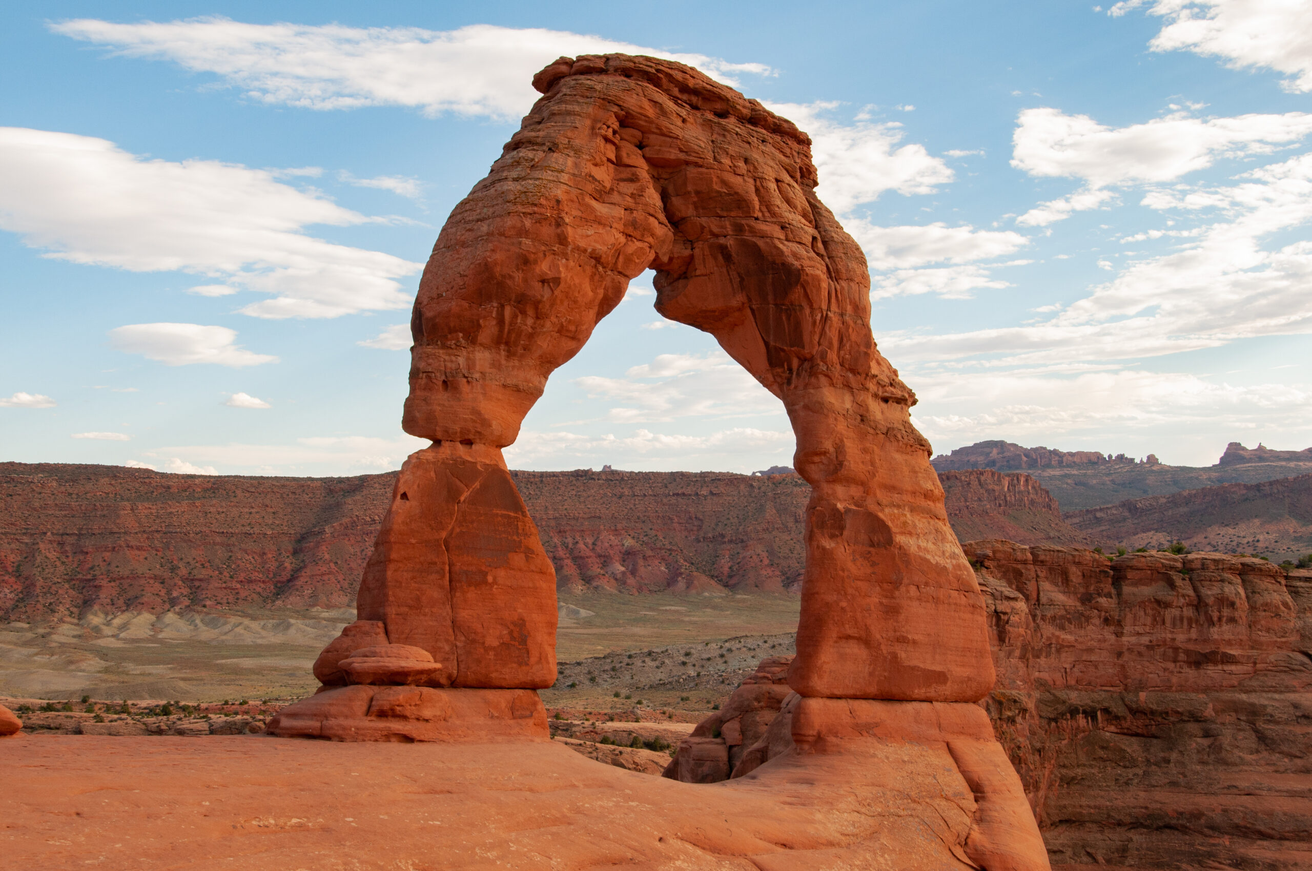 Red sandstone arch looming in a desert canyon under a bright blue sky with white clouds.