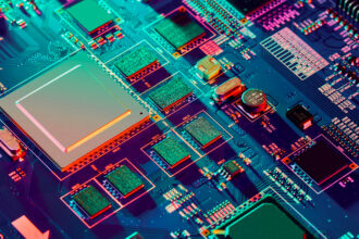 Close-up of a blue-green circuit board with chips, capacitors, and orange connectors illuminated in neon colors.