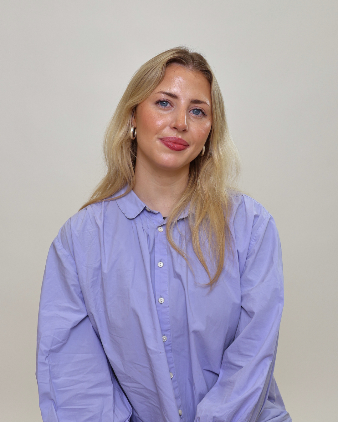 Portrait of a woman with long blonde hair, wearing a lavender button-down shirt, smiling softly against a neutral background.