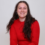 Enya Hughes - OHE Smiling woman with long dark wavy hair in a red sweater against a light gray background, portrait.