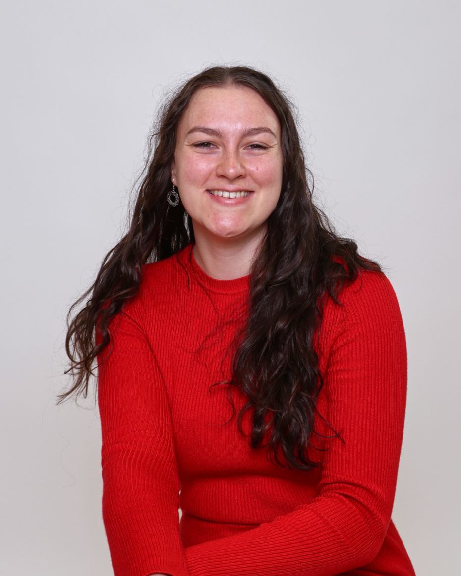 Smiling woman with long dark wavy hair in a red sweater against a light gray background, portrait.
