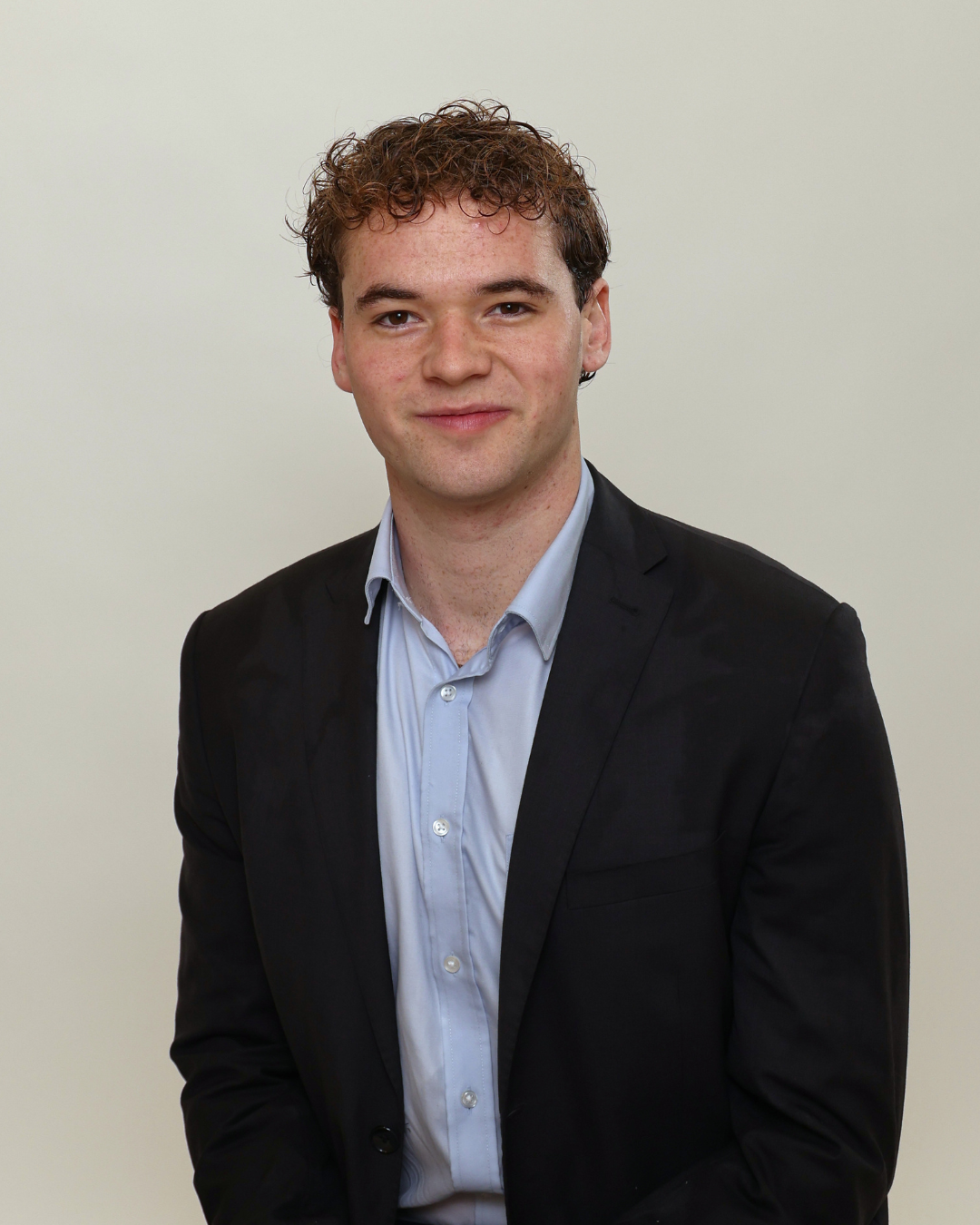Professional head-and-shoulders portrait of a young man with curly brown hair, wearing a black blazer and light blue dress shirt, smiling softly against a neutral background.
