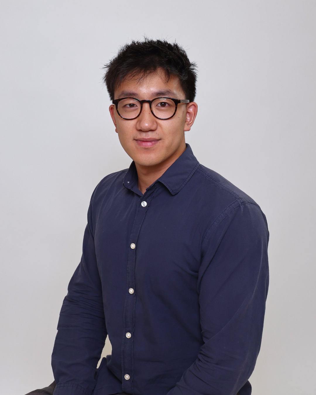 Professional head-and-shoulders portrait of a man wearing a navy button-down shirt and round glasses, looking at the camera with a slight smile in a neutral studio background.