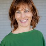 Julia Vitarello - OHE Smiling woman with auburn hair wearing a green sweater, posed outdoors against a gray brick wall behind her.