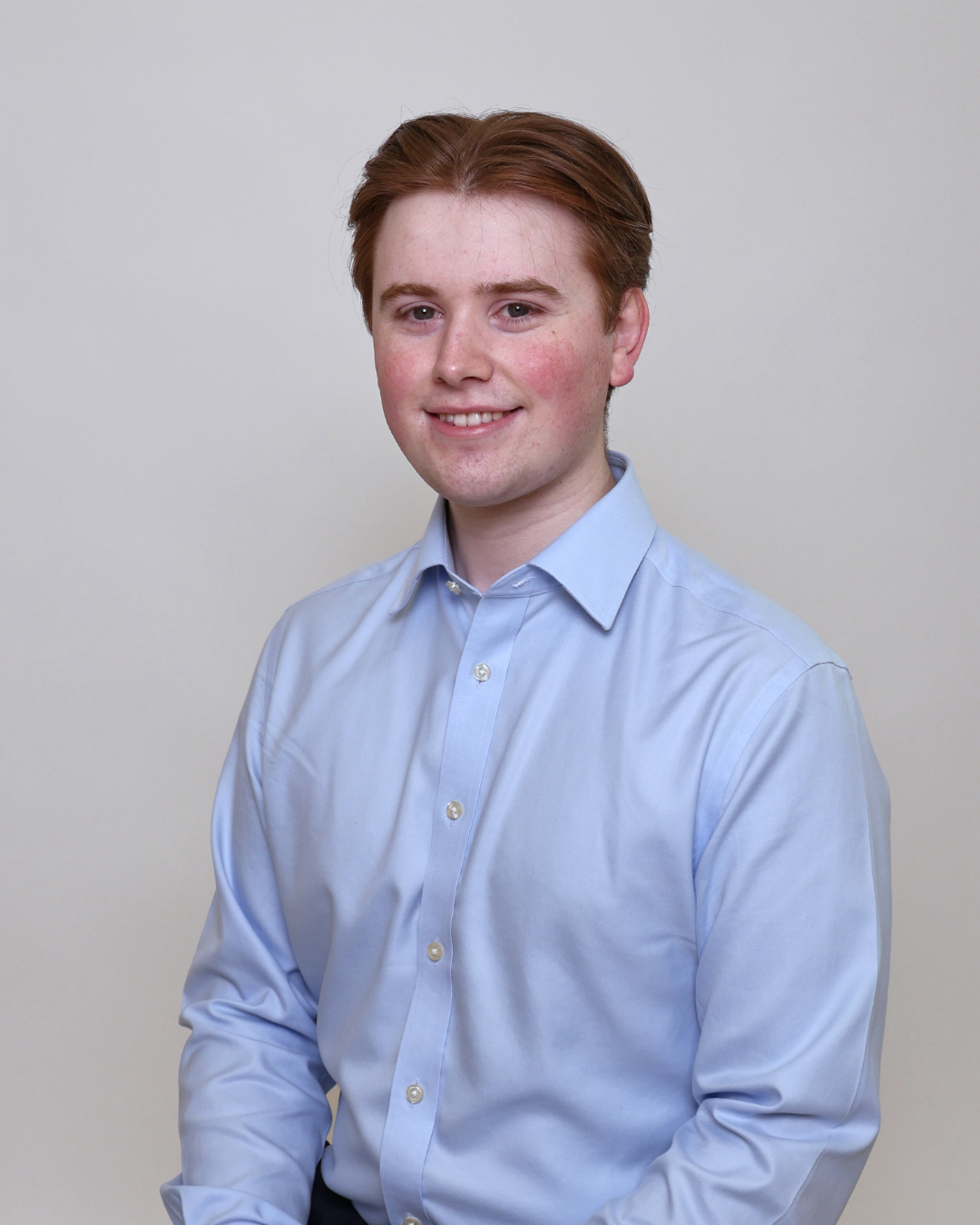 Portrait of a smiling young man with short red hair wearing a light blue dress shirt against a plain backdrop. (informational)